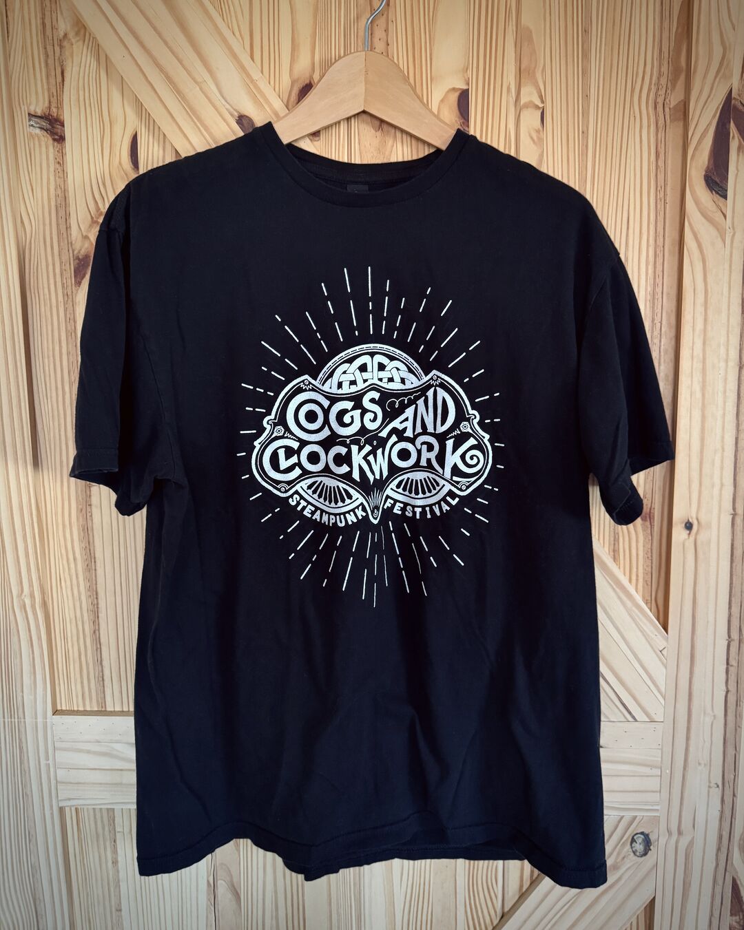 A black t-shirt on wooden hanger with a white print of "Cogs and Clockwork" festival logo featuring rays emanating from it.