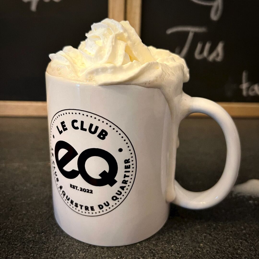 A cup of hot chocolate and whip cream in a Le Club EQ branded white mug on a dark table