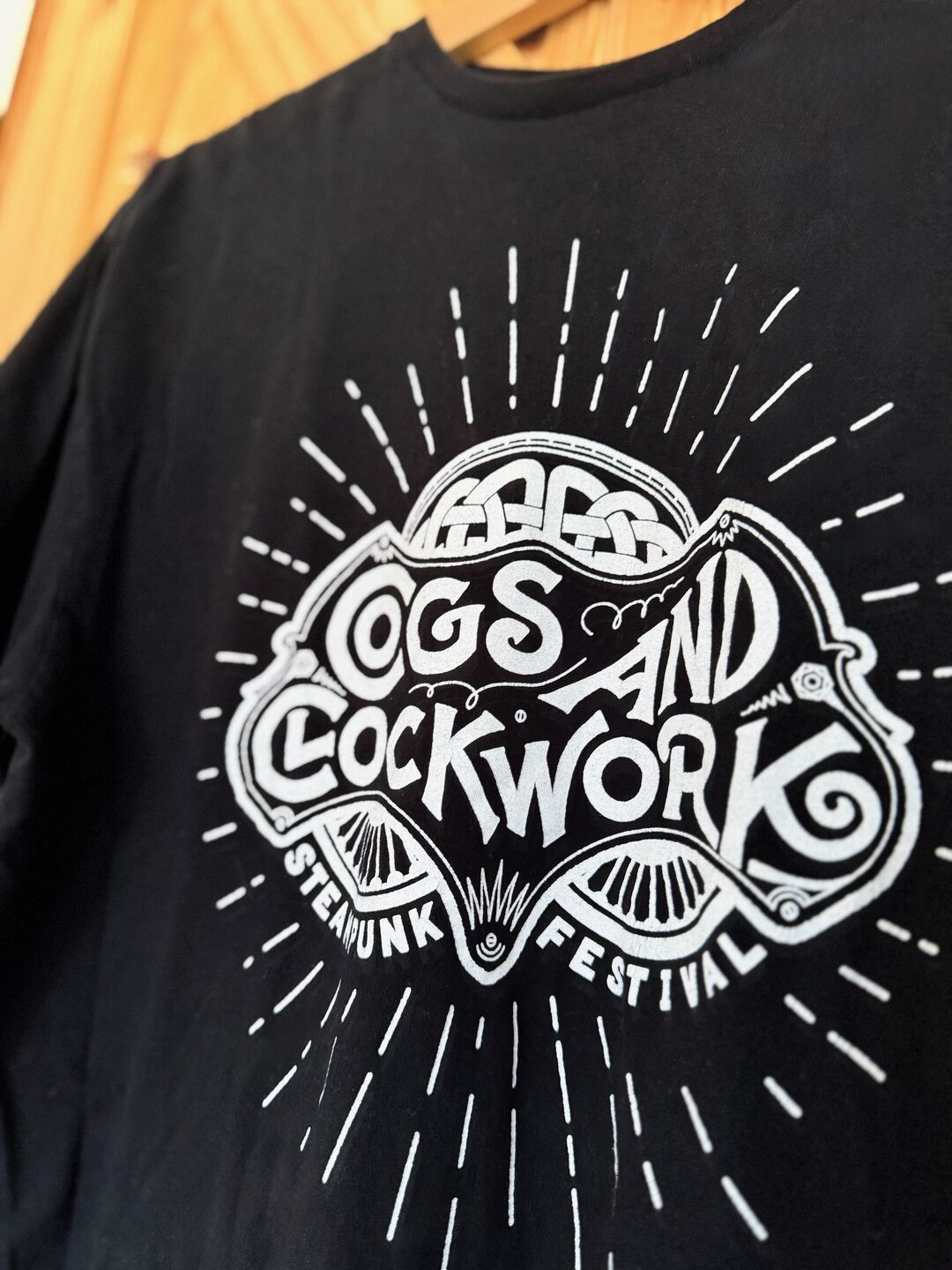 Detail of a black t-shirt on wooden hanger with a white printed "Cogs and Clockwork" festival logo featuring rays emanating from it.