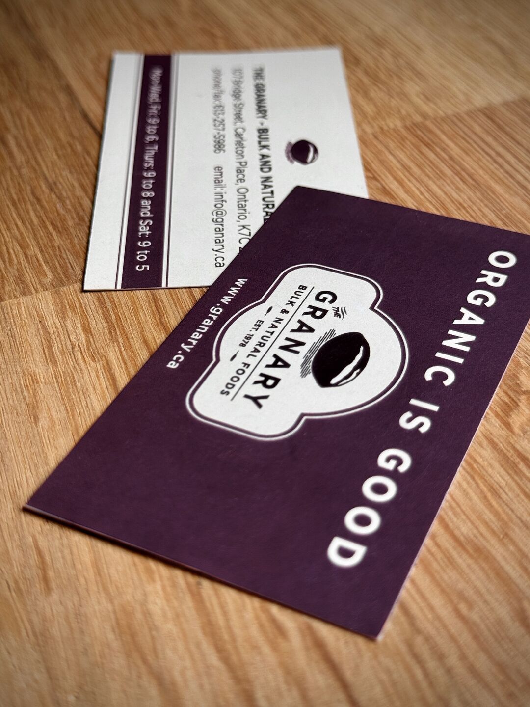 "The Granary" business card on wooden table. The tagline reads "Organic is Good" and includes a contact email address and phone number.