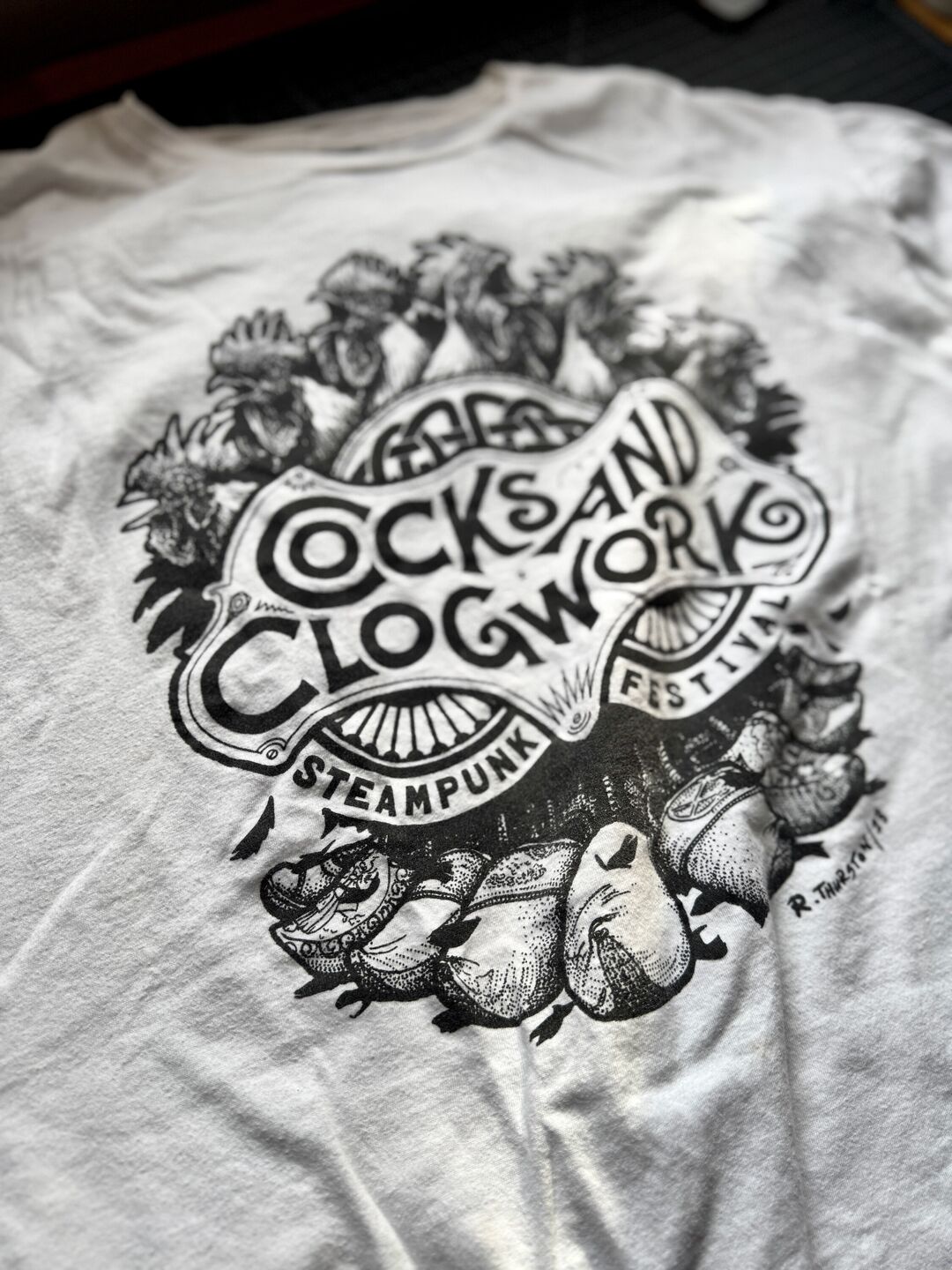 A white t-shirt with a graphic design featuring various illustrated crowing roosters wearing wooden shoes and text reading "Cocks and Clogwork" Steampunk Festival.