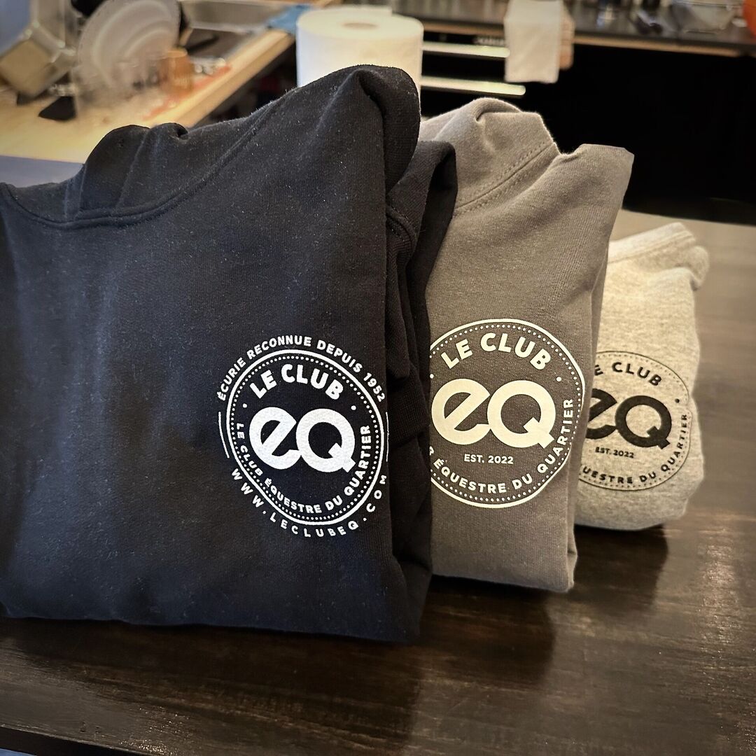 Three folded sweatshirts with Le Club EQ logos and text are displayed on a table.