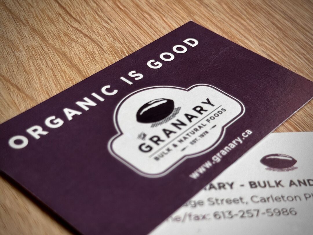 Detail of "The Granary" business cards on wooden table. The tagline reads "Organic is Good" and includes a contact email address and phone number.