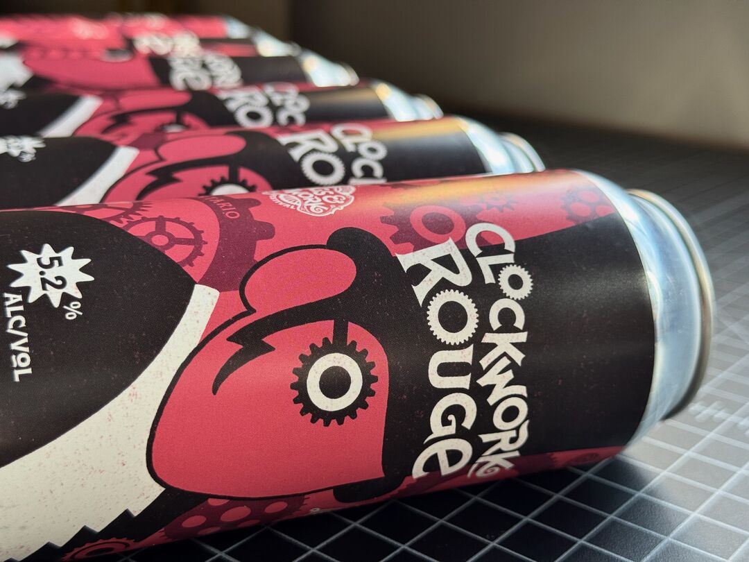 Clockwork Rouge beer cans in laid in a line on work table. Labels show a graphic styled ringmaster in front of exploded clock and cog mechanical parts on a salmon coloured field.