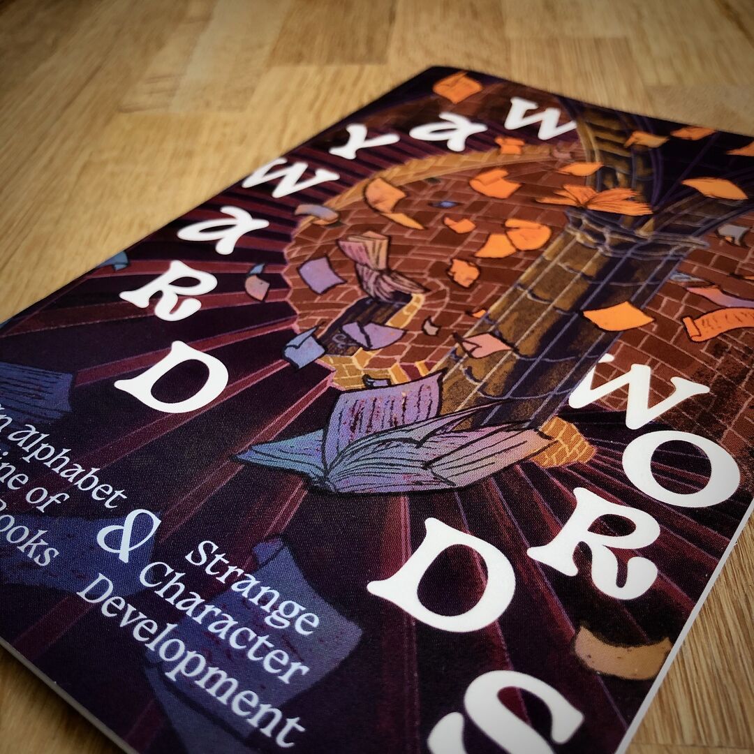 Close crop of "Wayward Words" zine cover depicting a chaotic jumble of pages, books and scrolls flying throughout a circular dungeon stairway is placed on a wooden surface.